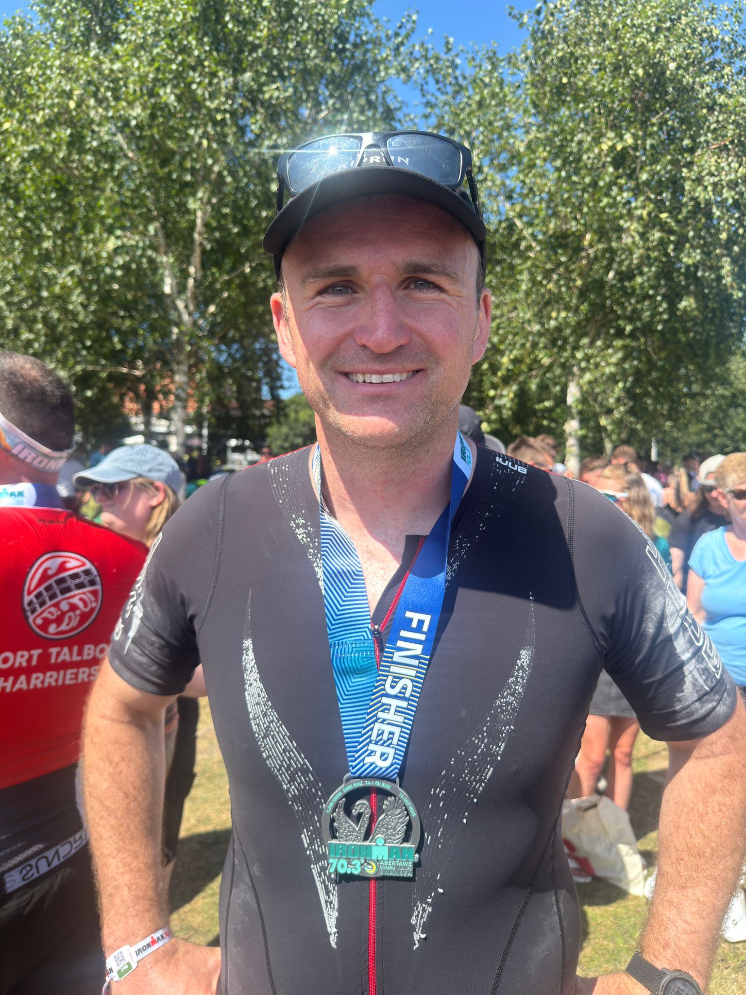 A smiling man wearing a black athletic suit and cap stands outdoors, proudly displaying a “Finisher” medal around his neck. People and trees are visible in the sunny background.