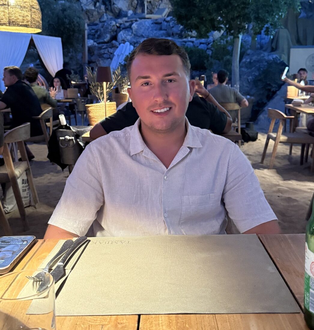 A man in a light shirt sits at a wooden table in an outdoor restaurant during the evening, surrounded by other diners. Bottles, glasses, and table settings are on the table. Warm lighting and greenery create a cozy atmosphere.