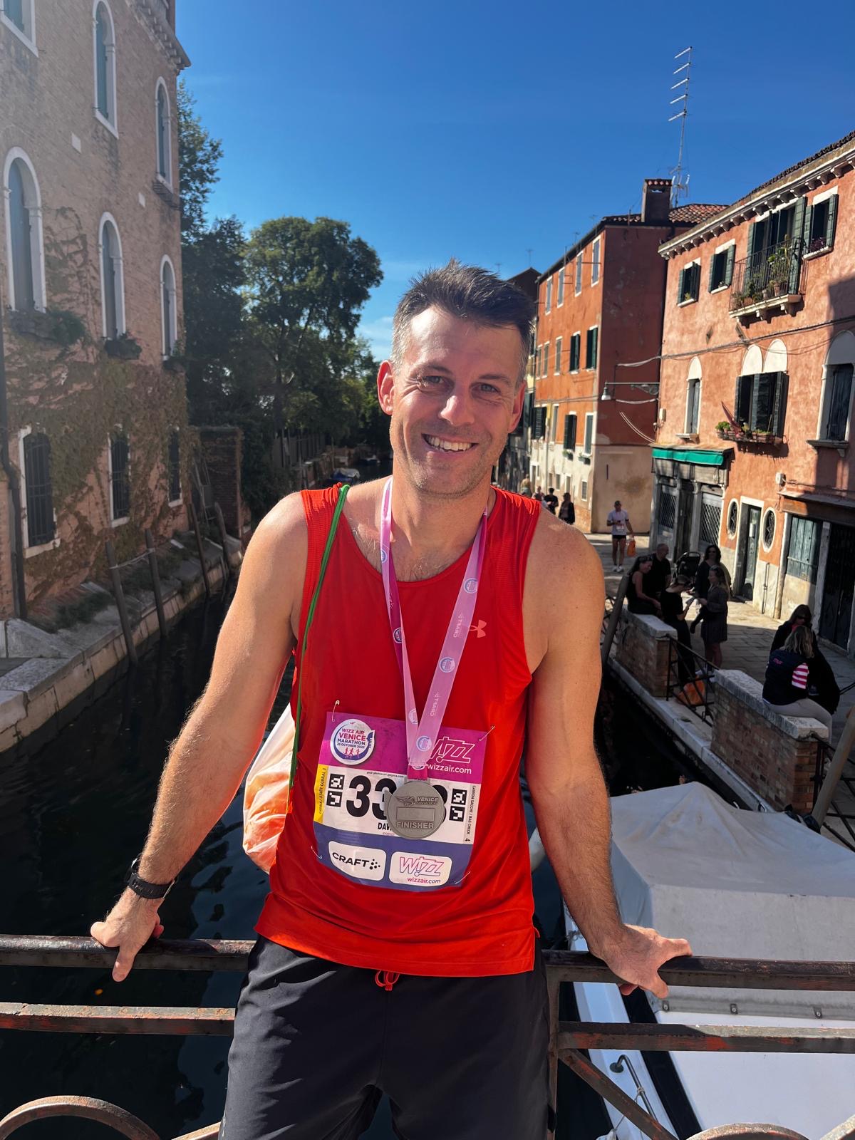 A smiling man in a red tank top with a race bib and medal stands on a bridge over a canal in a sunny, picturesque town with colorful buildings and people in the background.