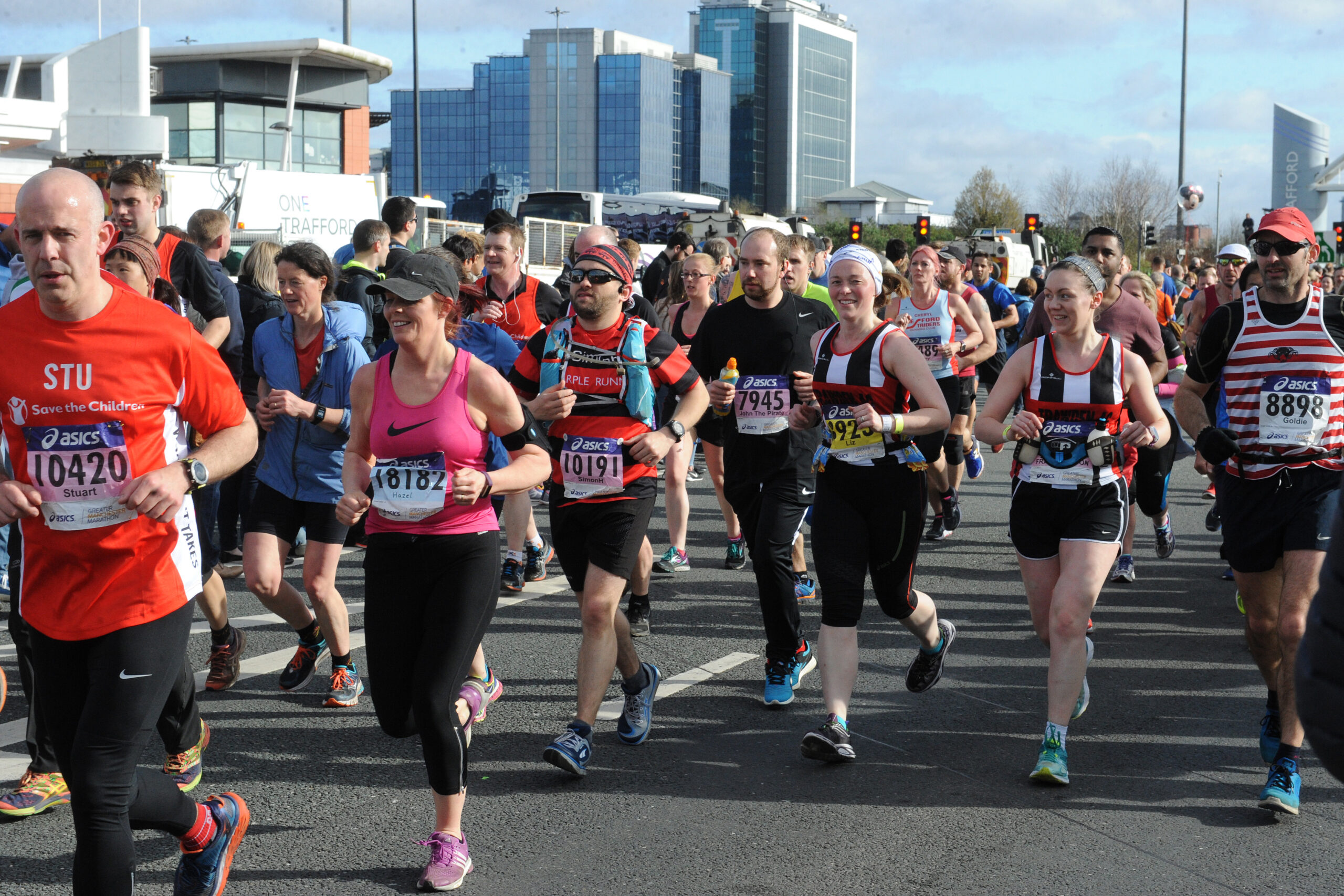 A group of marathon runners in athletic gear run on a city street, with tall buildings in the background and race bibs pinned to their shirts. Some runners are smiling while others look focused.