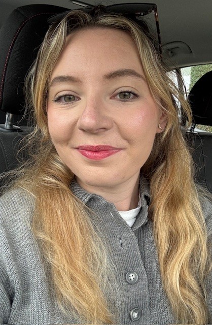 A woman with long blonde hair and light eyes is sitting in a car, smiling slightly at the camera. She is wearing a gray button-up sweater and sunglasses on her head.