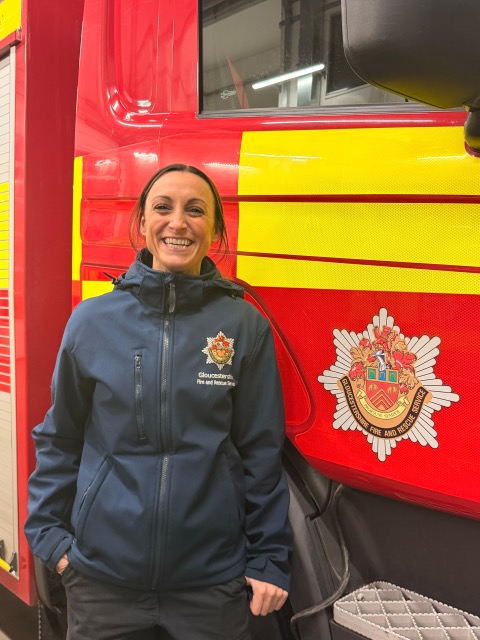 A smiling woman in a navy fire and rescue jacket stands next to a red fire engine with yellow stripes and an official fire and rescue service emblem.