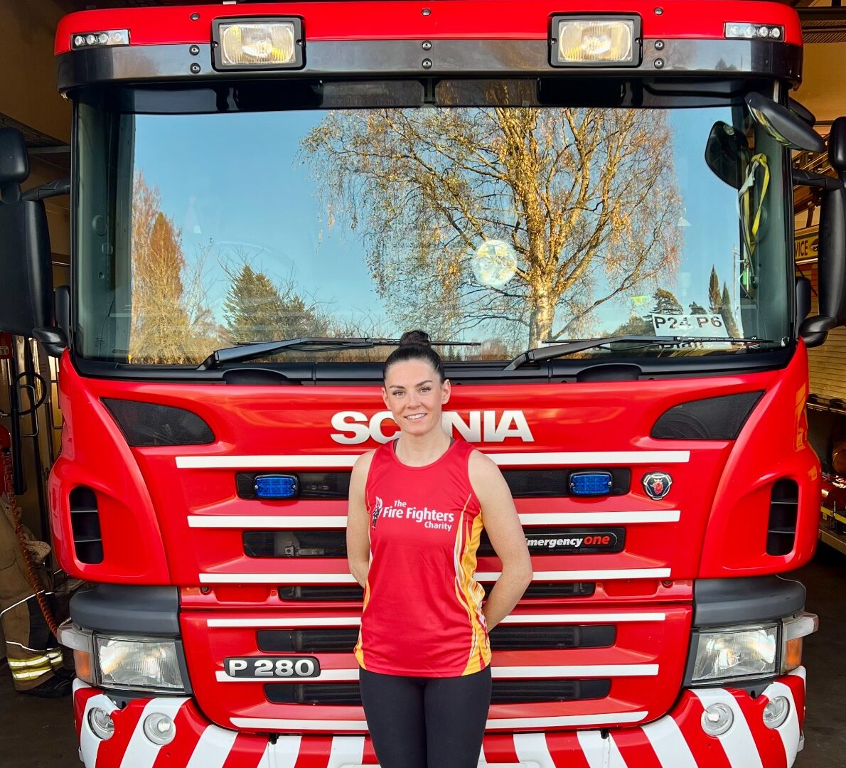 A woman in a red "Fire Fighters" tank top stands smiling in front of a large red Scania fire truck parked inside a fire station, with trees reflected in the truck’s windshield.