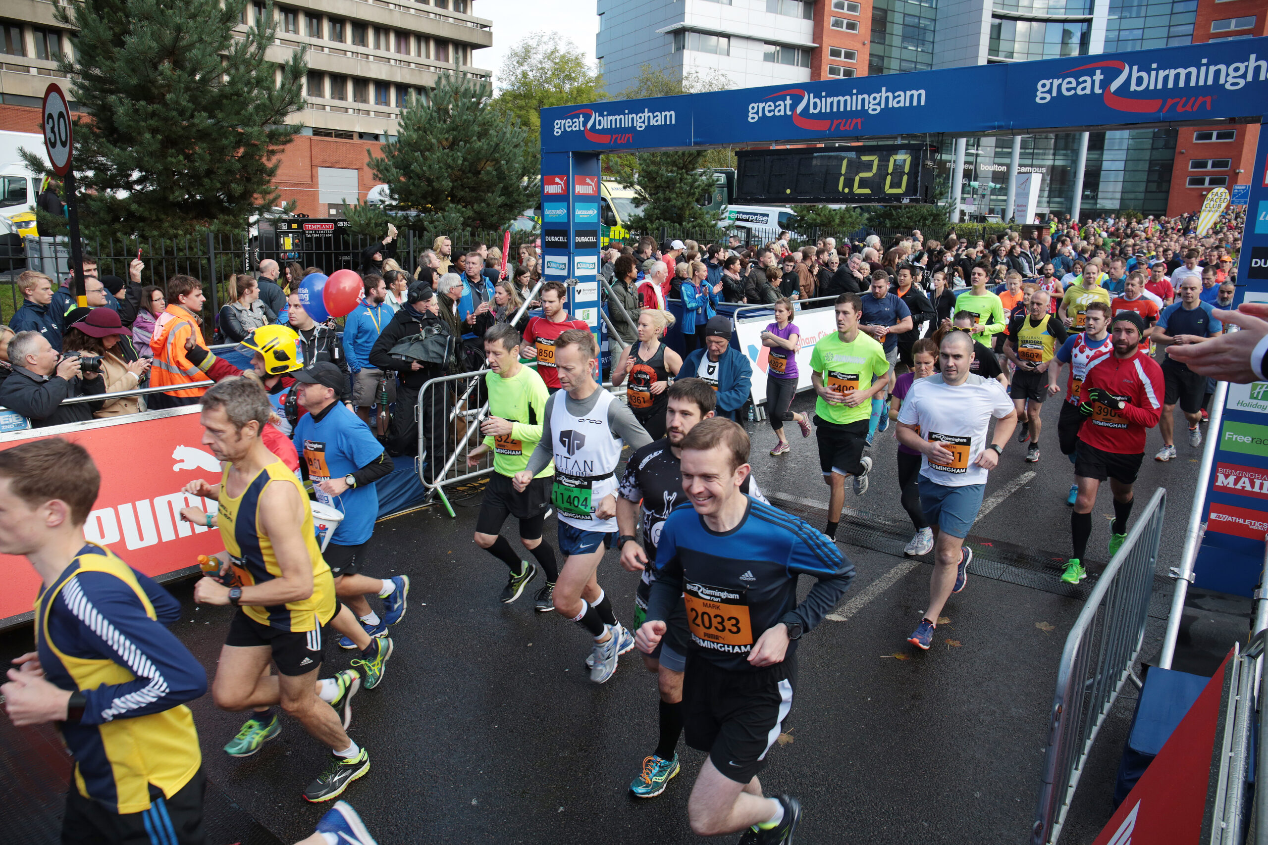 Runners wearing race numbers participate in the Great Birmingham Run, crossing the starting line with cheering crowds and event banners in the background on a cloudy day.