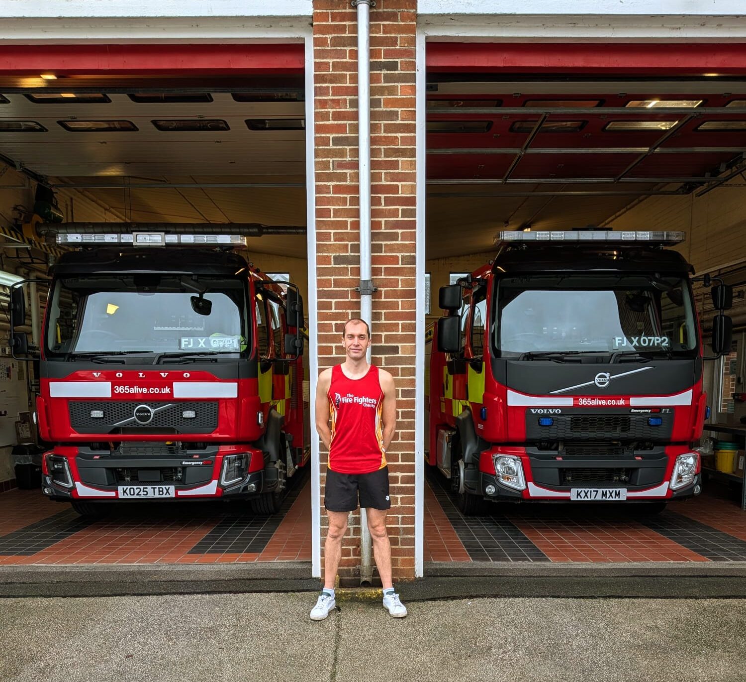 A person in athletic attire stands on wet pavement outside a fire station, positioned between two parked fire engines inside open garage bays. The fire station has a brick exterior.