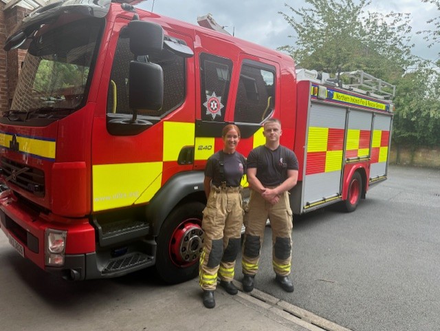 Two firefighters in uniform stand smiling in front of a red and yellow fire engine parked on a street, with trees and a building visible in the background.