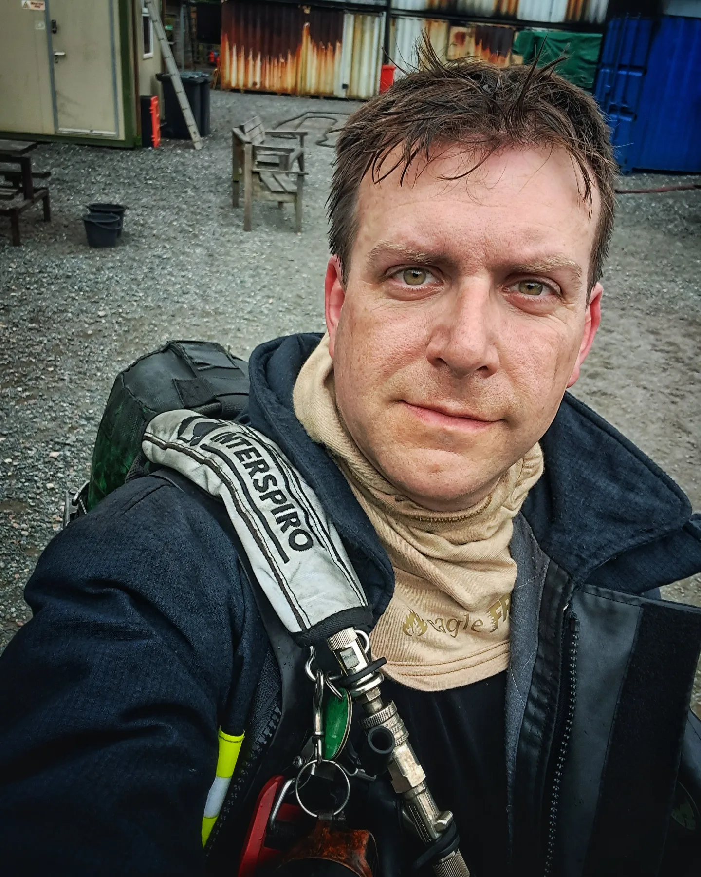 A man in firefighting gear takes a selfie outdoors, with a fire hose over his shoulder and training buildings in the background. He appears slightly sweaty and tired, suggesting recent physical exertion.