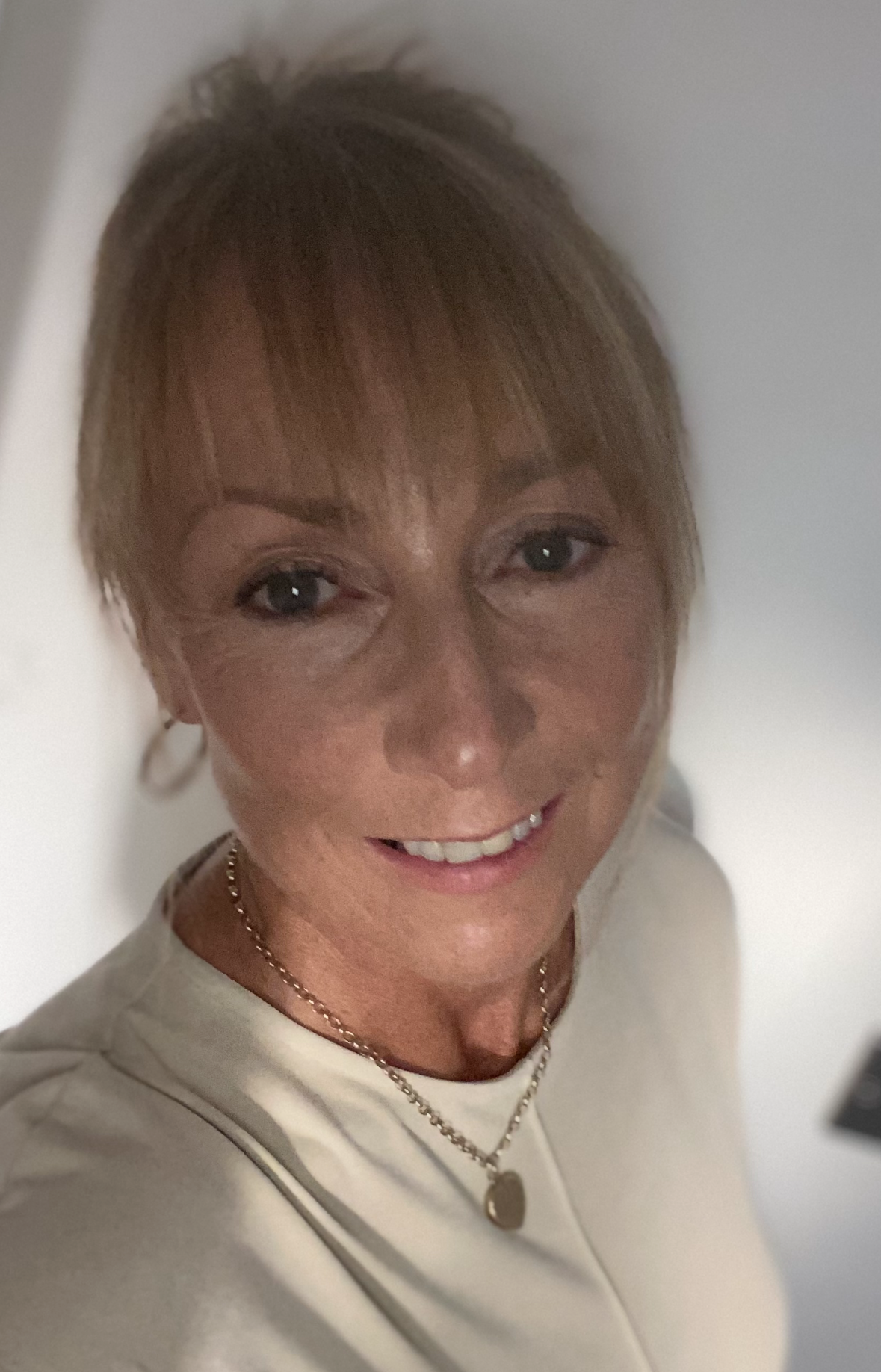 A woman with light brown hair and bangs, wearing a light-colored top and a gold necklace, smiles at the camera in a softly lit indoor setting.