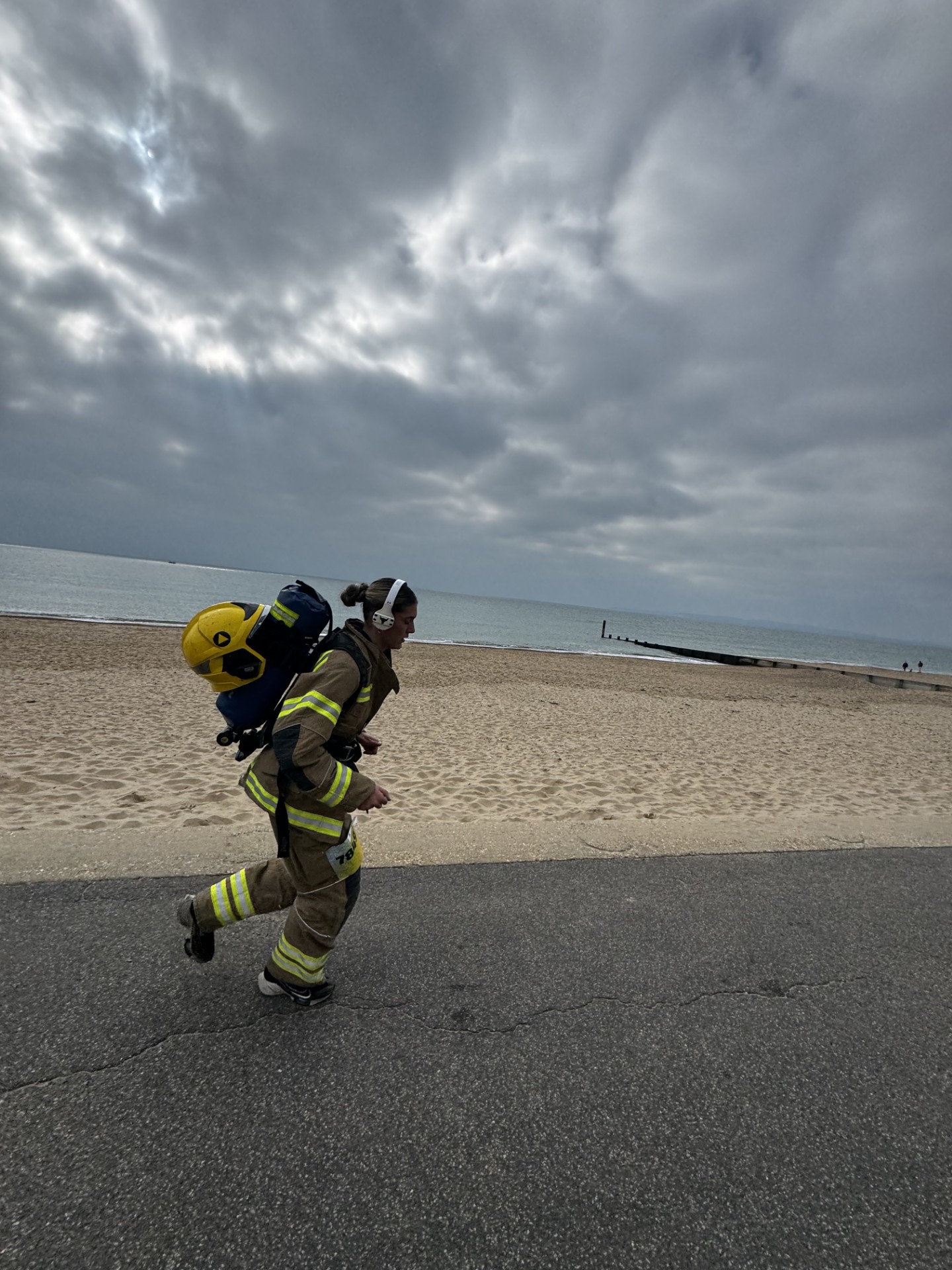 A firefighter in full gear runs on a paved path beside a sandy beach under a cloudy sky. The sea is visible in the background.