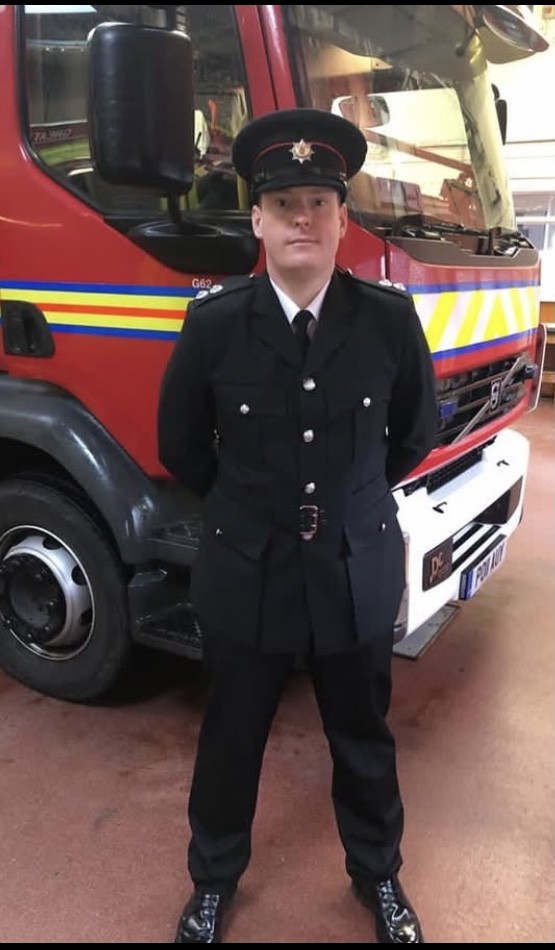 A firefighter in formal uniform stands with hands behind their back in front of a red fire engine inside a fire station.