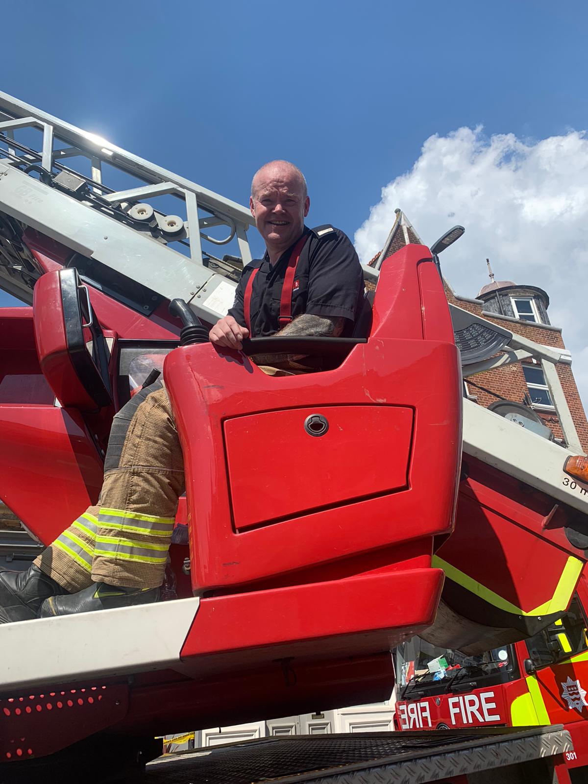 A firefighter in uniform sits in the cab of a red fire engine, smiling and looking at the camera. A blue sky with some clouds and part of a brick building are visible in the background.