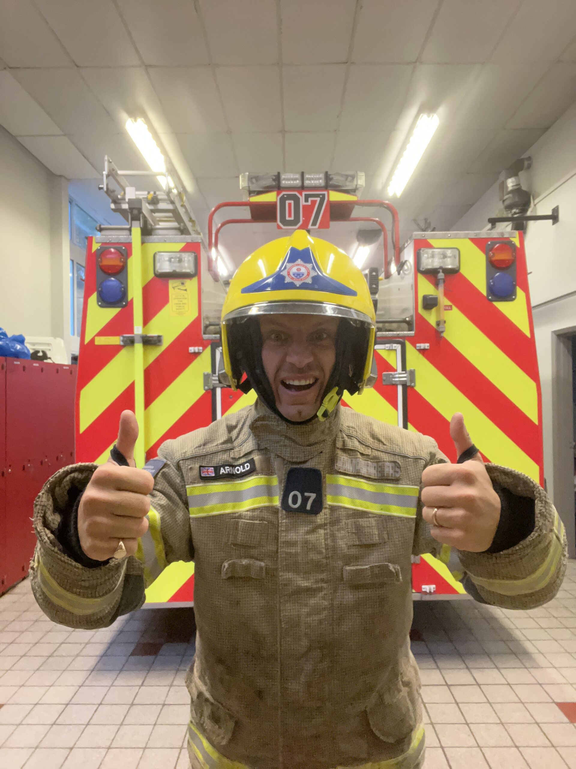 A firefighter in full gear and a yellow helmet stands indoors in front of a fire engine, smiling and giving two thumbs up. The fire engine has red and yellow chevron stripes and the number 07 on it.