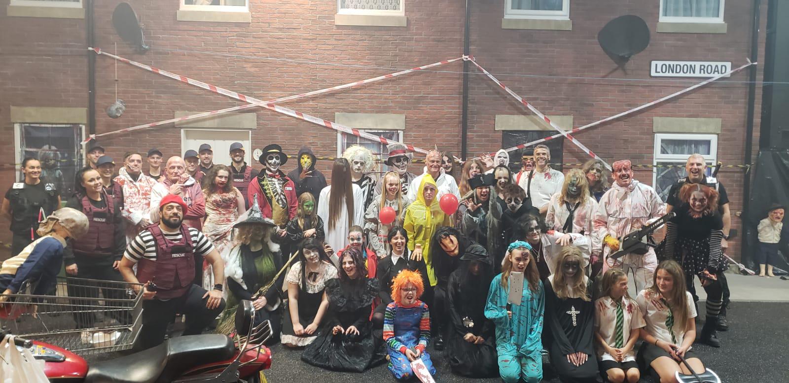A large group of people in diverse and elaborate Halloween costumes, including clowns, zombies, witches, and other spooky characters, pose for a photo on a street with a "London Road" sign and caution tape in the background.