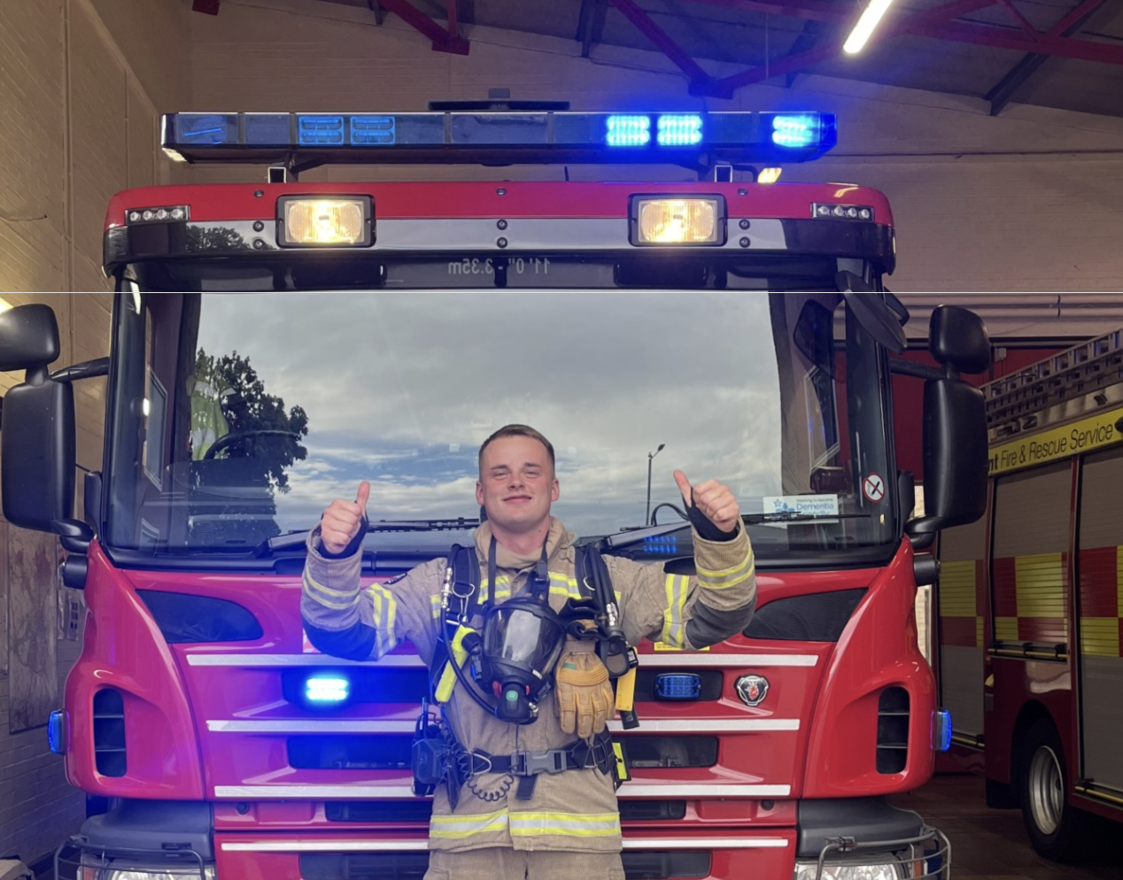A firefighter in full gear stands in front of a red fire engine with blue lights flashing, giving two thumbs up and smiling. The fire station is visible behind the vehicle.