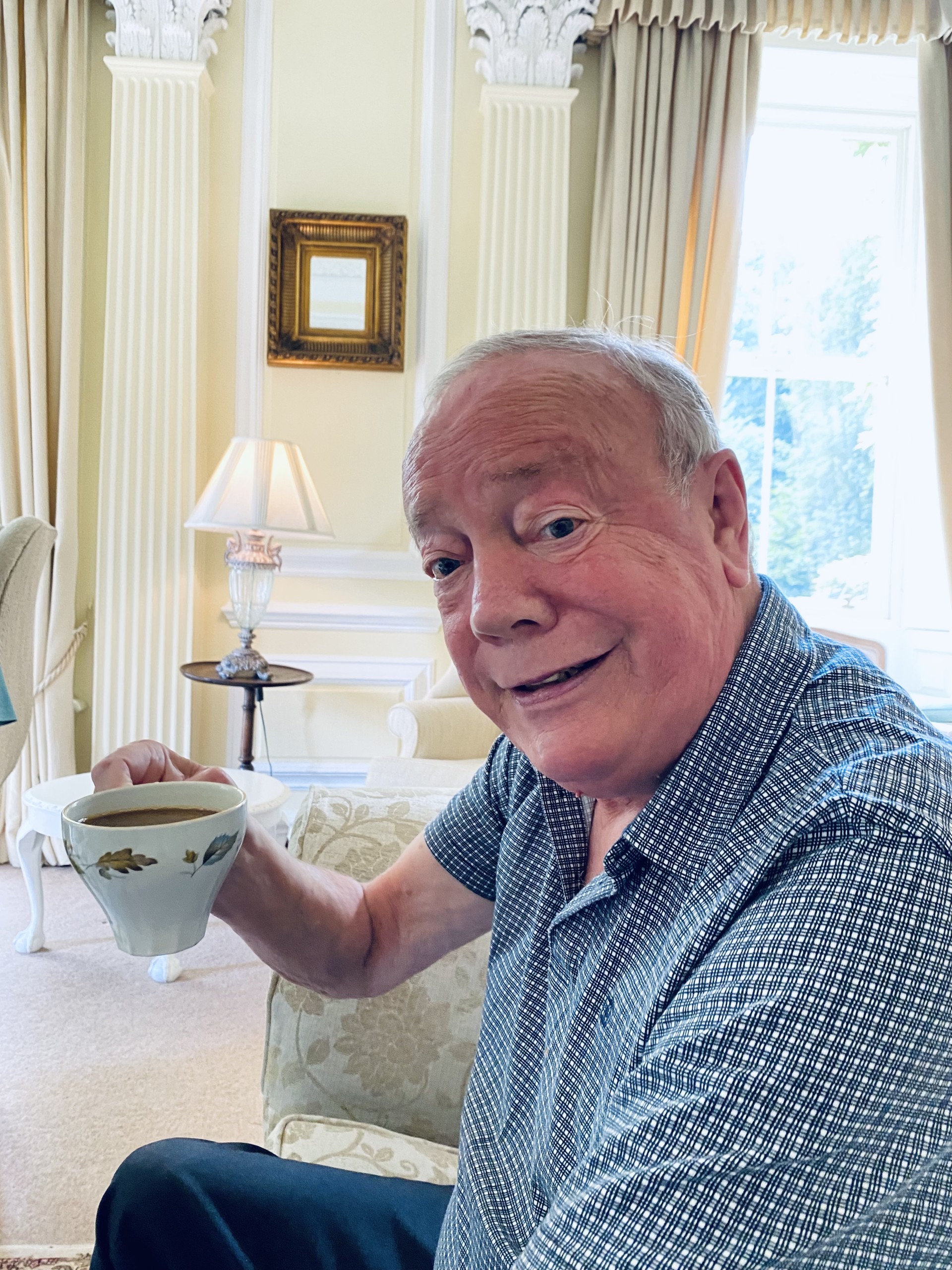 An older man with short gray hair smiles while holding a teacup in an elegant, well-lit living room with cream-colored decor and large windows.