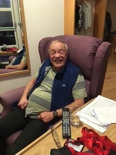 An older man with glasses and a blue vest sits smiling in a mauve armchair by a table with papers, a TV remote, and a glass of water. A coat rack and an open window are visible in the background.
