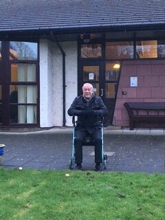An elderly man sits on a walker outside a building, smiling at the camera. The building has large windows, a door, and a bench nearby. The grass and pavement are wet, suggesting it has recently rained.