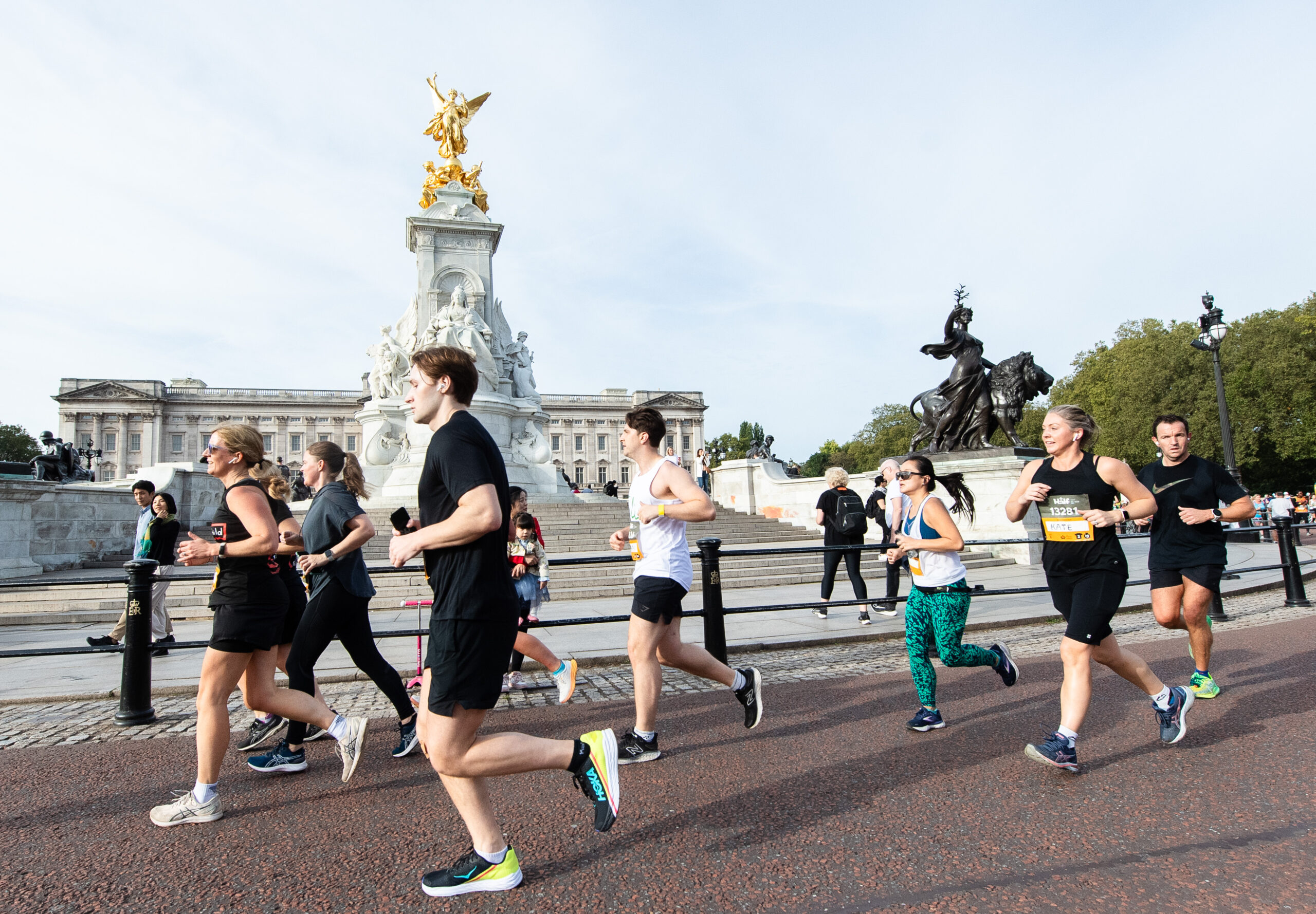 Runners taking part in a race pass in front of the Victoria Memorial near Buckingham Palace on a sunny day, with spectators and historic statues visible in the background.