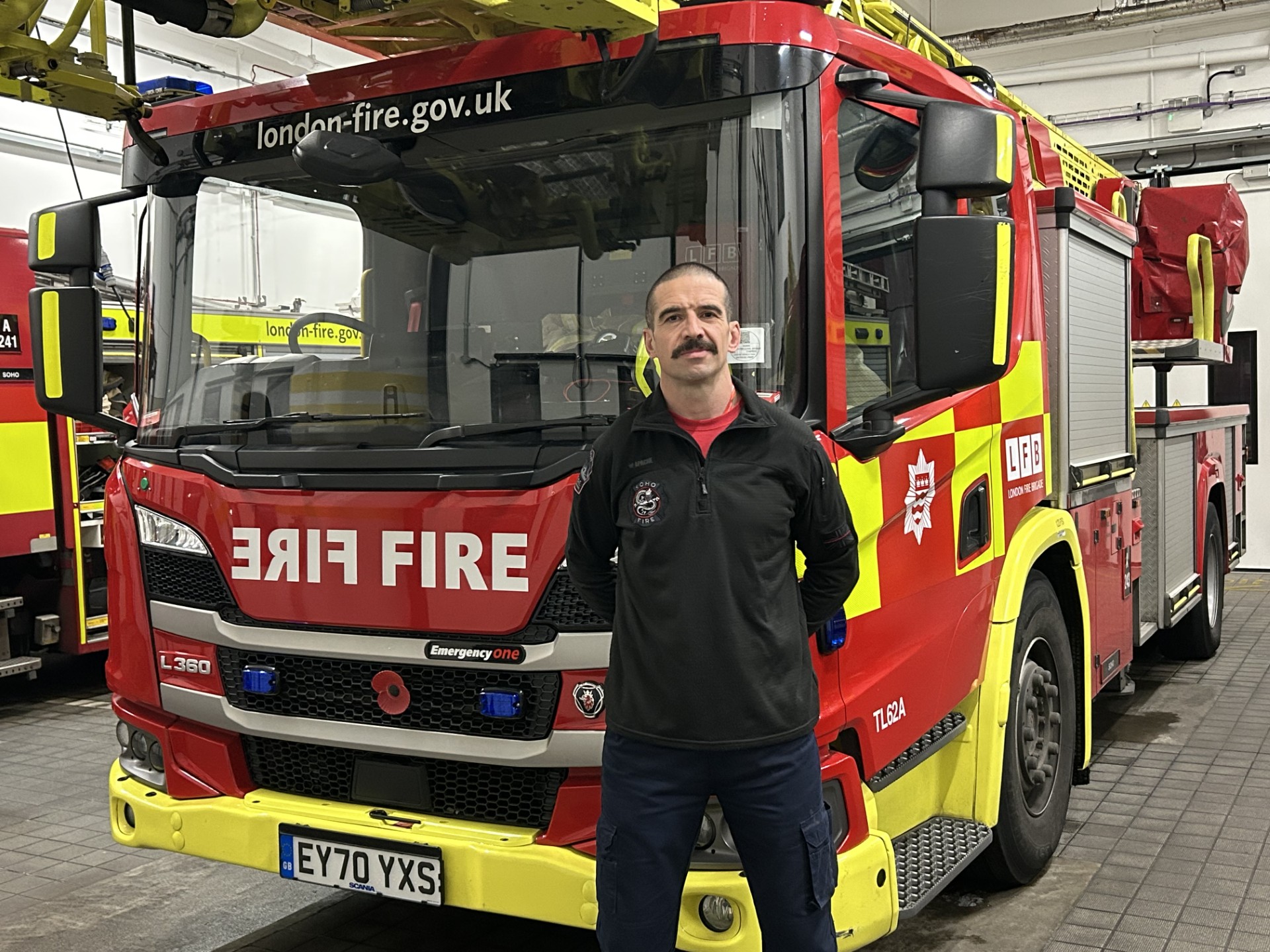 A firefighter with a mustache stands in front of a red London Fire Brigade fire truck inside a fire station. The truck has yellow accents and emergency equipment on top.