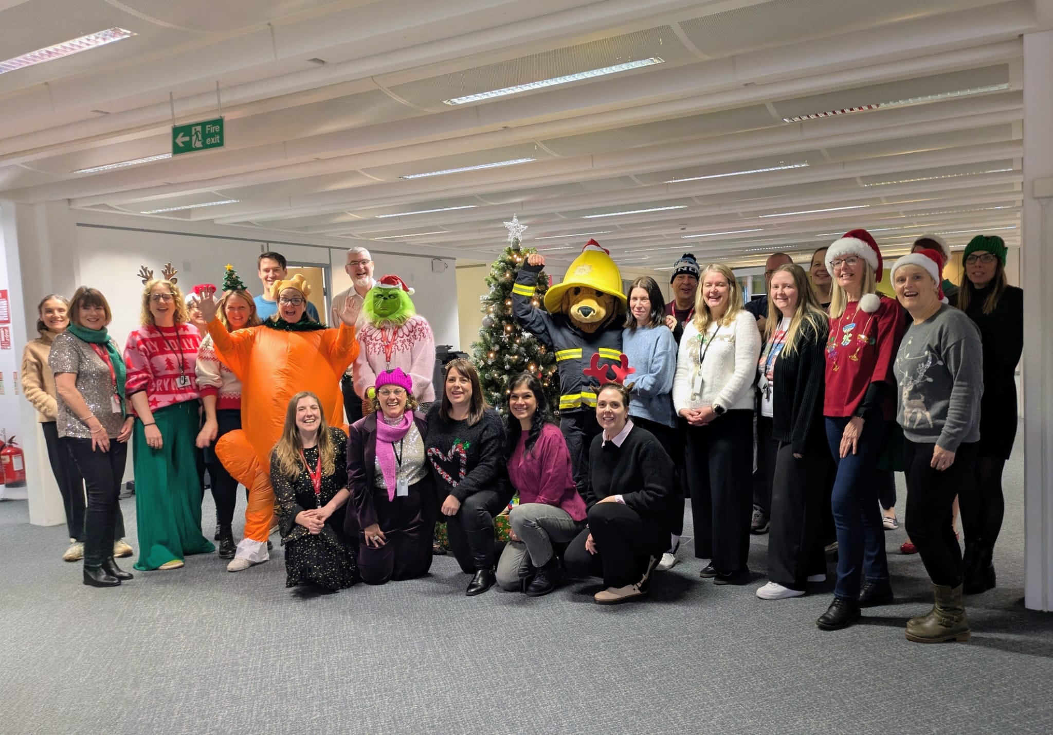 A group of people posing around a decorated Christmas tree in an office, some wearing festive outfits, hats, or costumes, including a bear and an orange inflatable suit. They are all smiling and celebrating together.