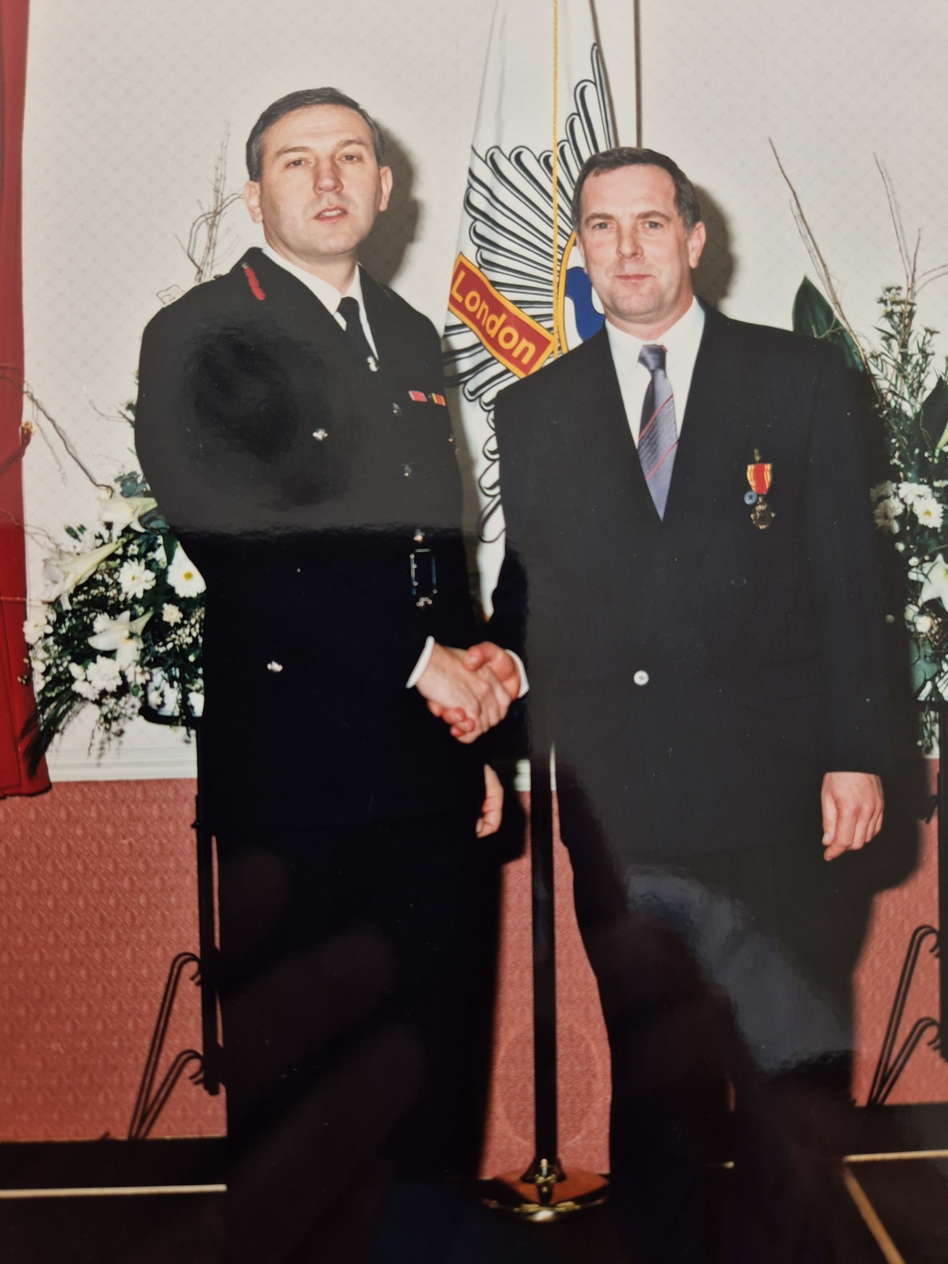 Two men in formal attire shake hands in front of a flag and floral arrangements. One man wears a uniform, the other a suit with a medal. The setting suggests an official event or ceremony.
