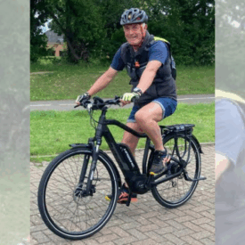 A smiling man wearing a helmet, shorts, and a backpack is riding a black bicycle on a paved path with grass and trees in the background.