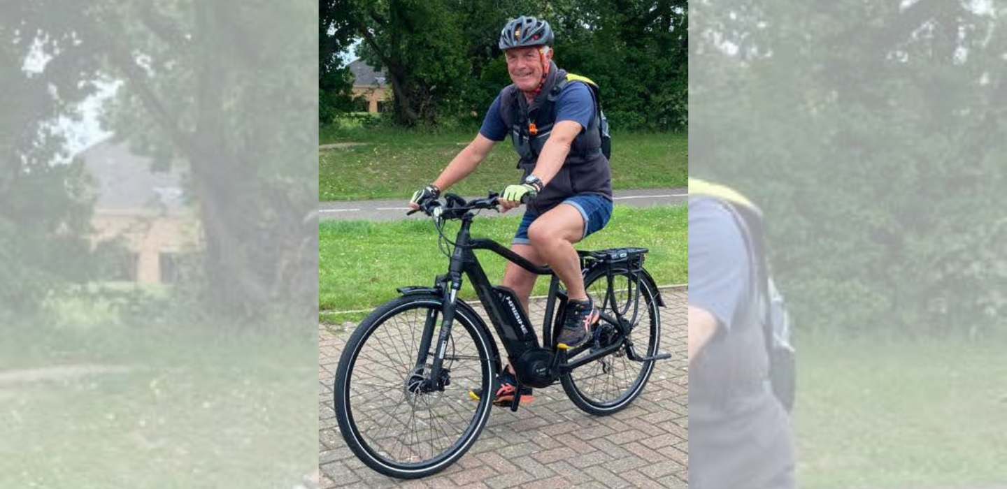 A smiling man wearing a helmet, shorts, and a backpack is riding a black bicycle on a paved path with grass and trees in the background.