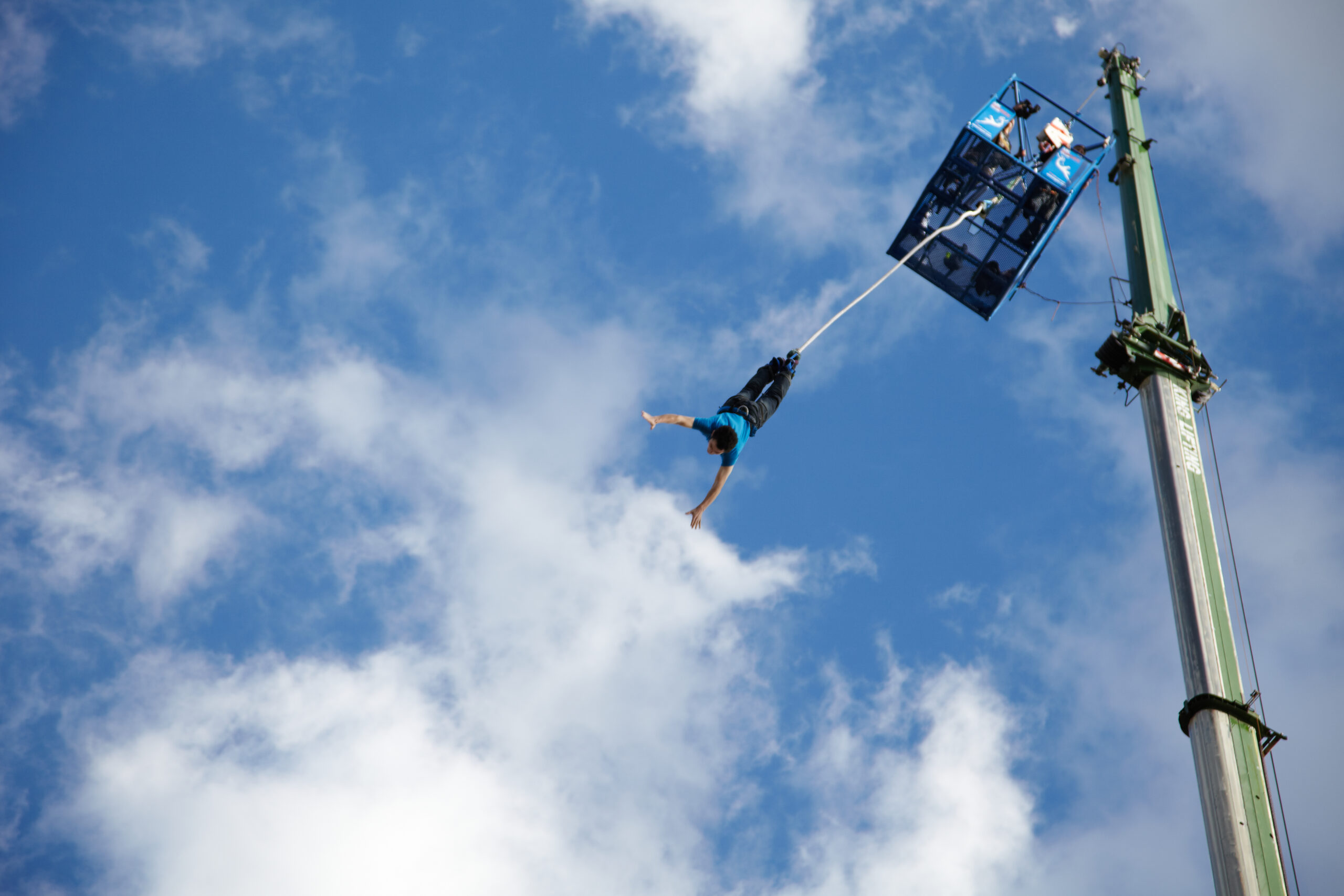 A person is bungee jumping from a high platform attached to a crane, arms spread wide, against a backdrop of blue sky and scattered clouds.