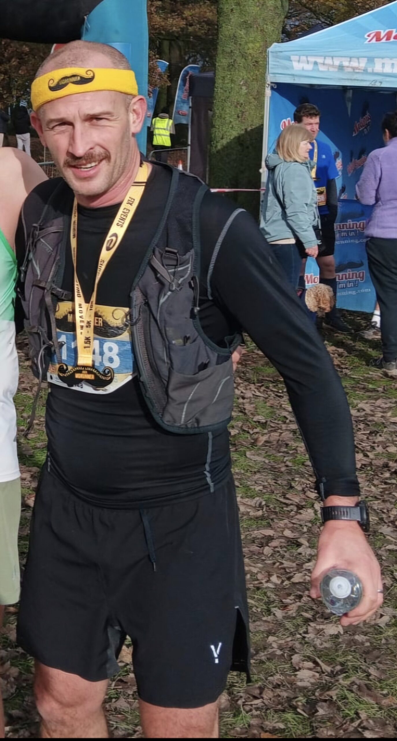 A man wearing a yellow headband with a mustache design, a race bib, and a medal stands outdoors on a leafy ground, holding a water bottle. He is dressed in black athletic gear and a running vest. People and a blue tent are in the background.