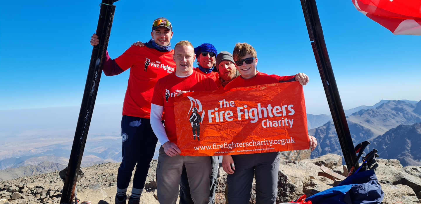 Four people in red shirts stand on a mountain summit, holding a banner that reads “The Fire Fighters Charity.” Rugged mountain peaks are visible in the background under a clear blue sky.