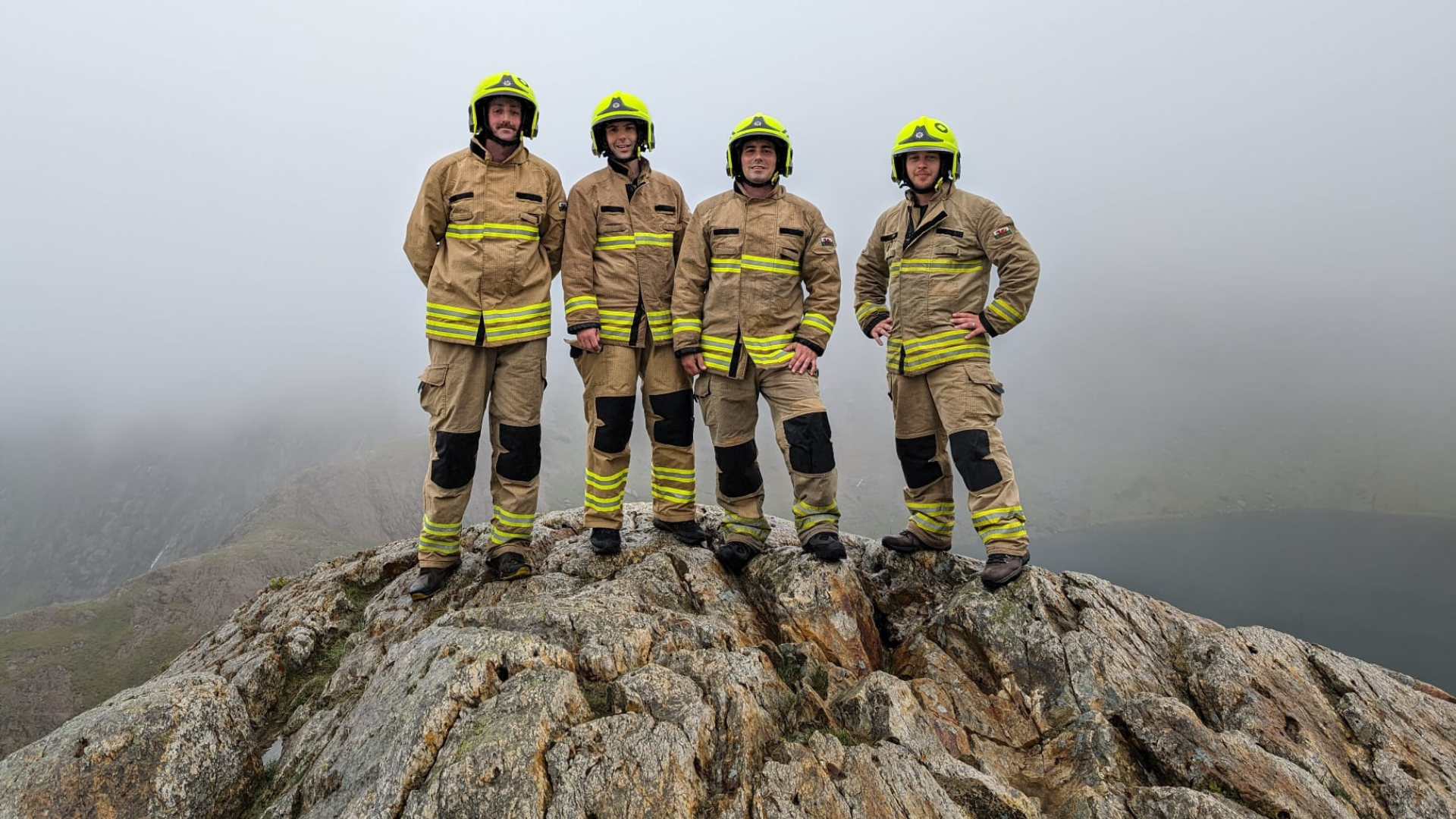 Four firefighters in uniform and helmets stand together on a rocky mountain peak with misty, cloudy skies in the background.