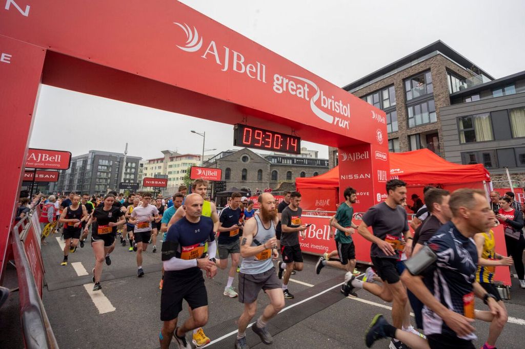 Runners start the AJ Bell Great Bristol Run, passing under a large red banner with a digital timer. The street is lined with buildings and spectators, and the atmosphere is energetic and lively.