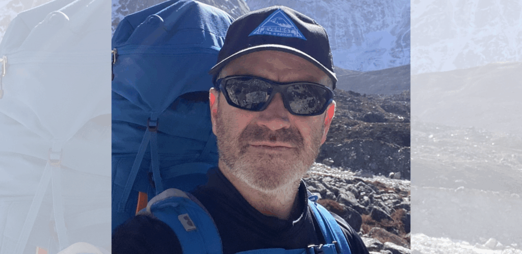 A man wearing sunglasses, a black cap, and a blue backpack takes a selfie outdoors in a rocky, mountainous area with snow-covered peaks in the background, under bright sunlight.