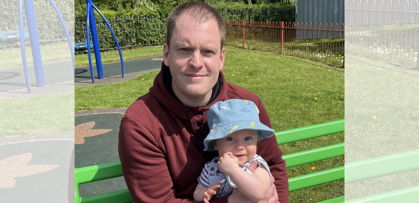 A man in a maroon hoodie sits on a green bench in a park, holding a smiling baby wearing a blue hat. Playground equipment and grass are visible in the background on a sunny day.