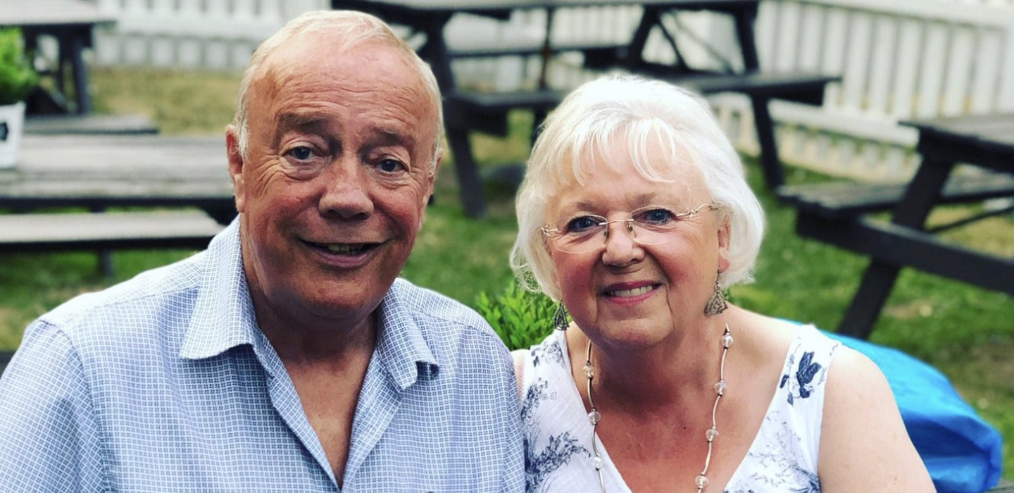 An older man and woman sit closely together outdoors, smiling at the camera. They are surrounded by wooden picnic tables and a white fence in the background on a sunny day.