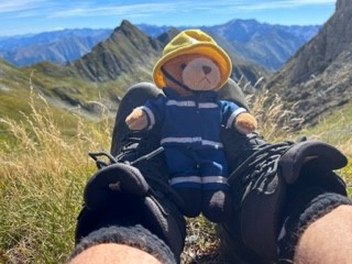 A person wearing hiking boots sits in grassy mountains, holding a small teddy bear dressed in a blue outfit and yellow hat, with rugged peaks and a clear blue sky in the background.