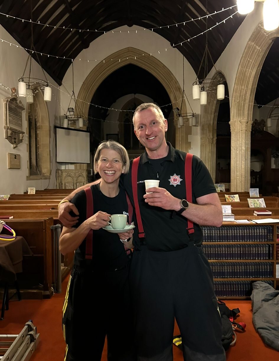 Two smiling firefighters in uniform stand together inside a church, each holding a cup. They wear black shirts and red suspenders, and the church's arched architecture and hanging lights are visible in the background.