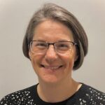 A woman with short brown and gray hair, wearing glasses and a black sweater with decorative sparkles, smiles while standing in front of a light gray background.