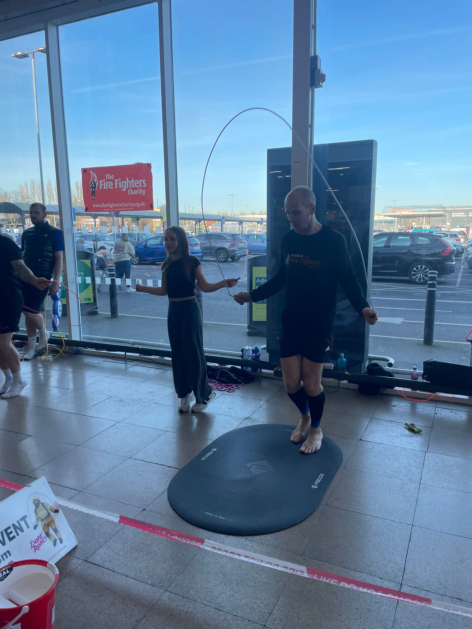 A man in athletic wear skips rope on a black mat indoors while a woman stands nearby holding a stopwatch. The scene is set near large windows with cars visible in the parking lot outside.