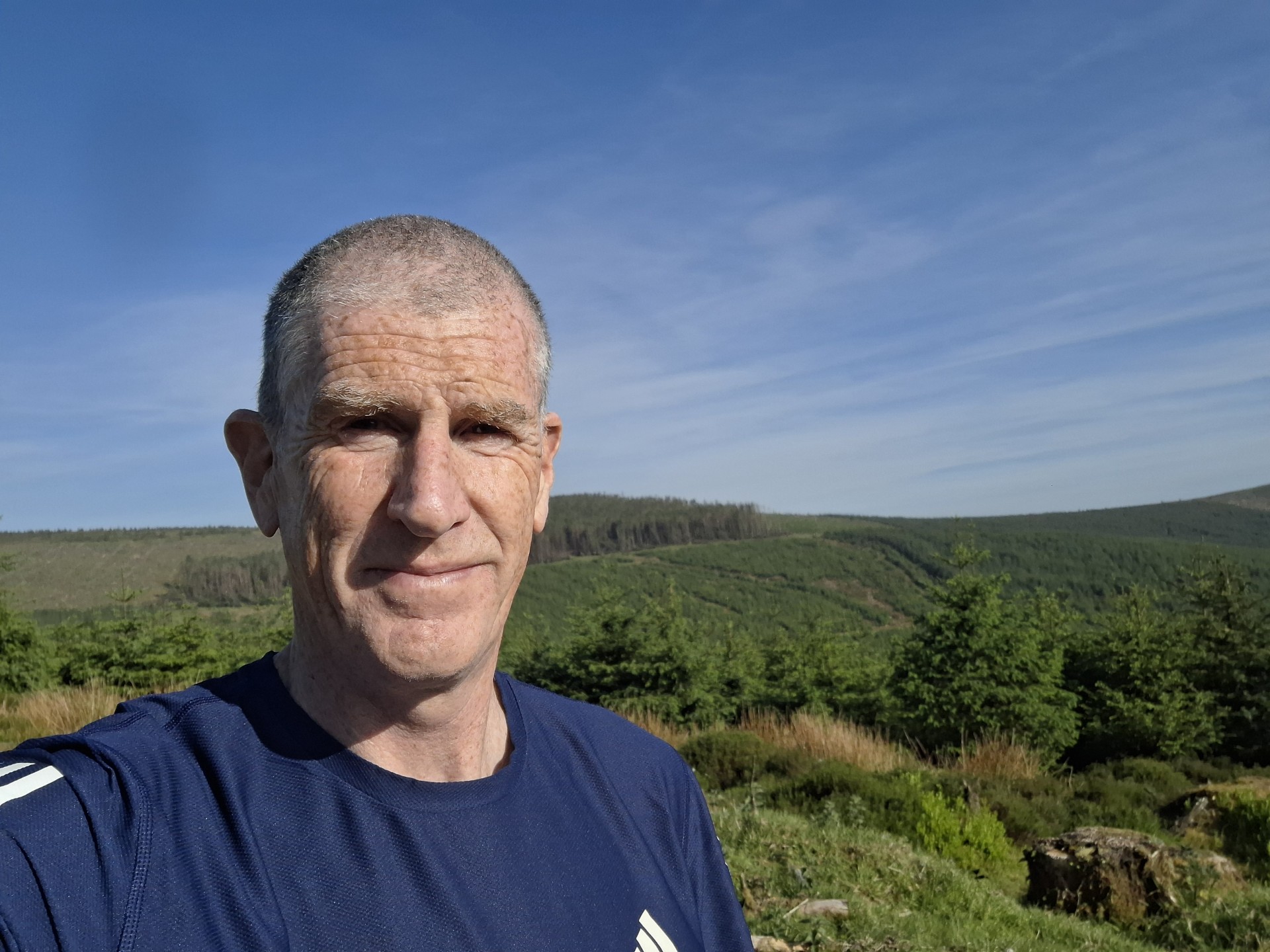 A man in a blue athletic shirt stands outdoors, smiling with hills, green trees, and a blue sky in the background.