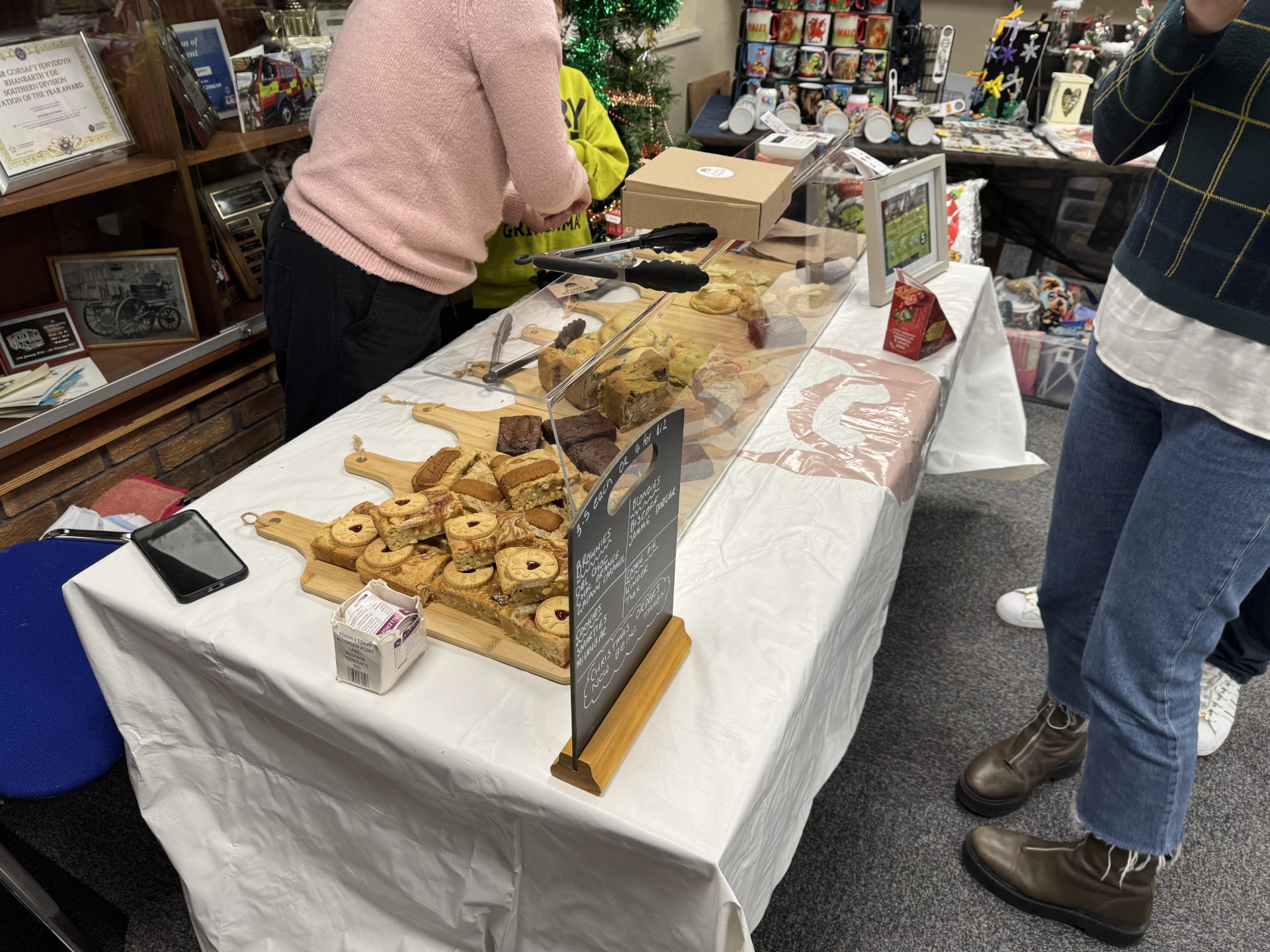 A table covered with a white cloth displays various pastries and baked goods on wooden boards. People stand nearby, and a plastic barrier separates the food from customers. A small sign and a phone are on the table.