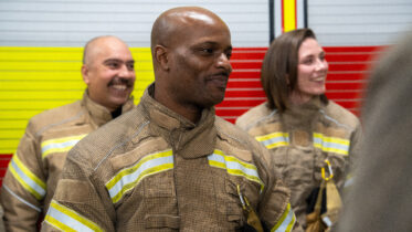 Three firefighters in uniform stand together, smiling. The background features a wall with red, yellow, and gray horizontal stripes. The focus is on the firefighter in the center.