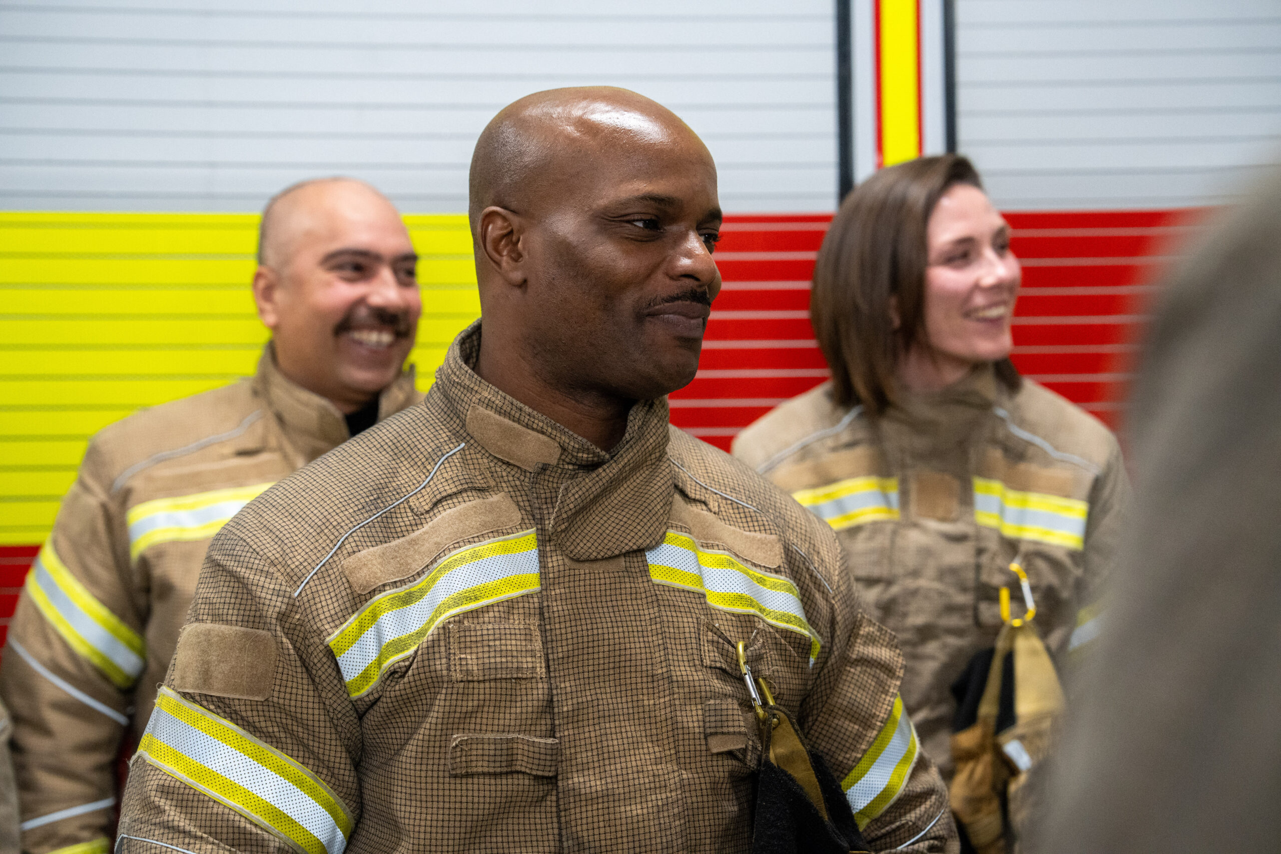 Three firefighters in uniform stand together, smiling. The background features a wall with red, yellow, and gray horizontal stripes. The focus is on the firefighter in the center.