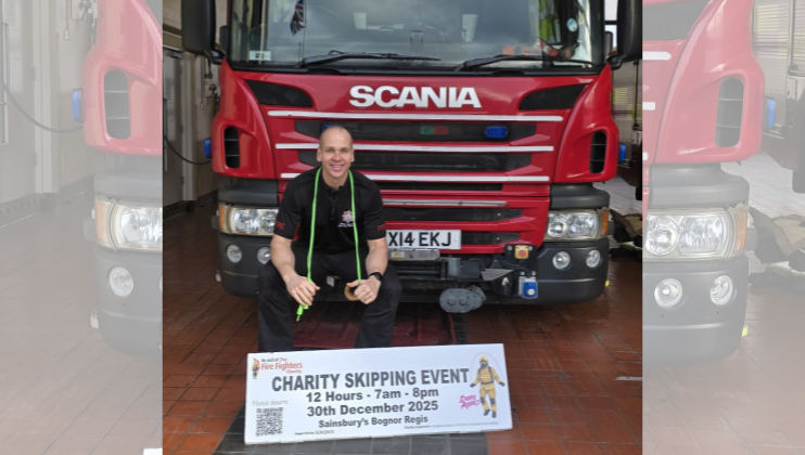 A man smiles while sitting in front of a red Scania fire truck. In front of him, a sign reads “Charity Skipping Event, 12 Hours - 7am-8pm, 30th December 2025, Sainsbury’s Bognor Regis.”.