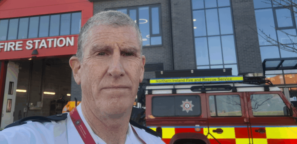 A man in a fire service uniform stands outside a fire station, with a red Northern Ireland Fire and Rescue Service vehicle parked behind him. The fire station building is visible in the background.