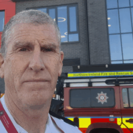 A man in a fire service uniform stands outside a fire station, with a red Northern Ireland Fire and Rescue Service vehicle parked behind him. The fire station building is visible in the background.