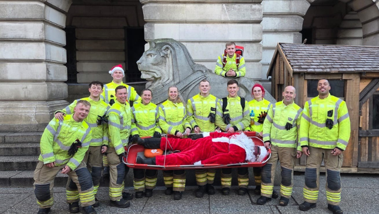 A group of firefighters in yellow uniforms pose and smile outside a building. They are holding a stretcher with a person dressed as Santa Claus lying on it. One firefighter wears a Santa hat.