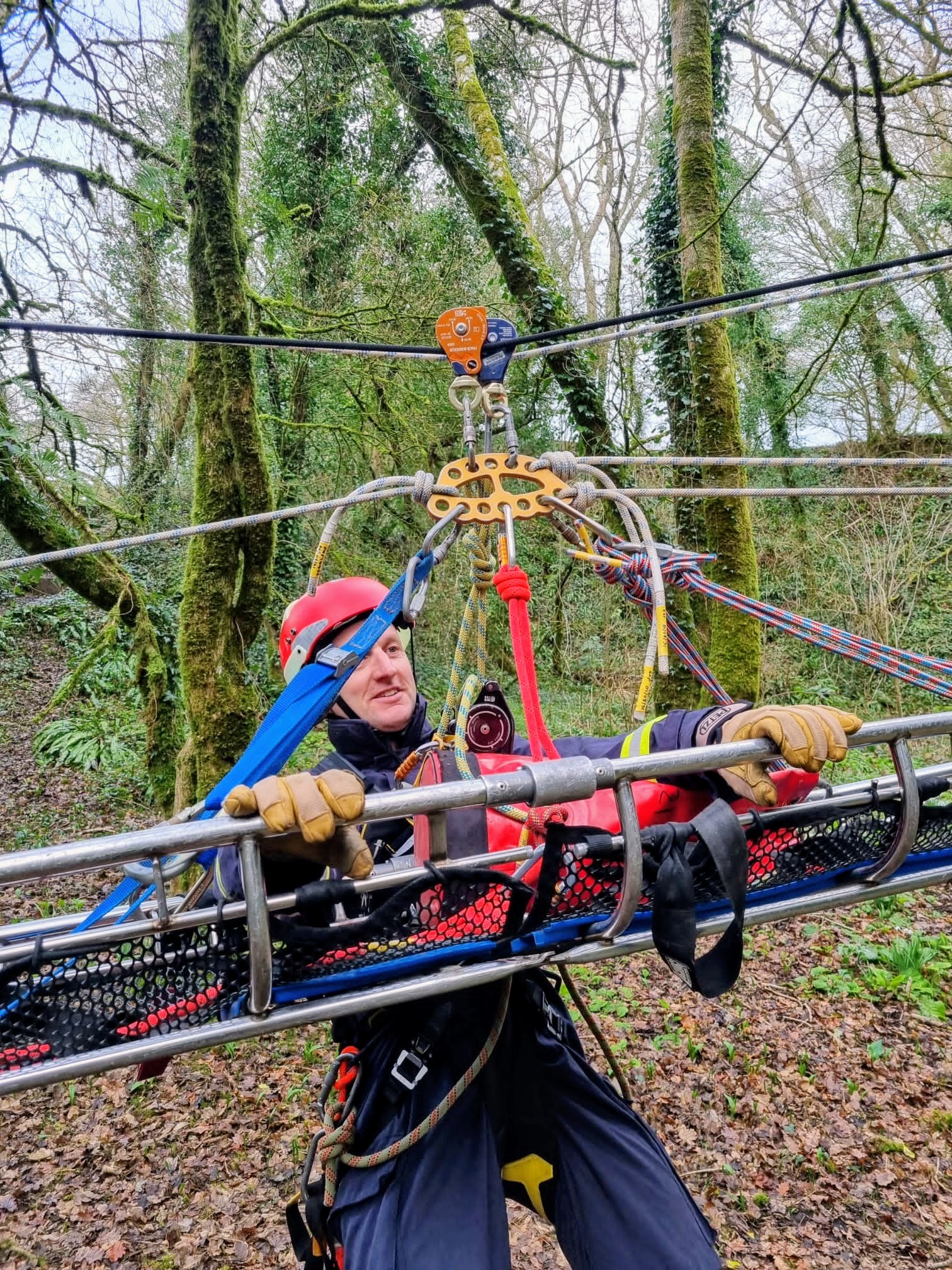 A person in protective gear and helmet operates a pulley system to transport a stretcher through a wooded area, suggesting a rescue training exercise. The scene is surrounded by trees and various rescue ropes.