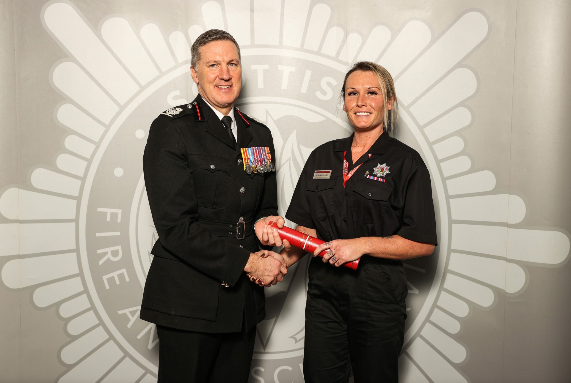 A man in a decorated uniform shakes hands with a woman in a black uniform holding a red certificate. They are standing in front of a large Scottish Fire and Rescue Service emblem.