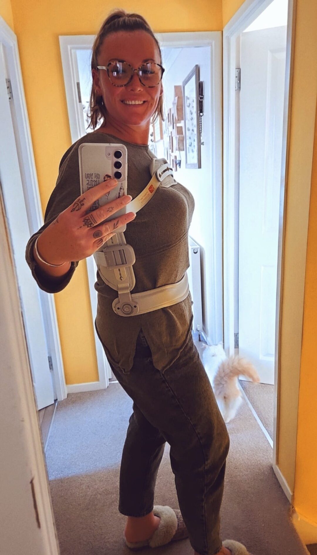 A smiling person wearing glasses and an orthopedic back brace takes a mirror selfie in a hallway. They are dressed in casual clothes and slippers, and a fluffy cat’s tail is visible in the background.
